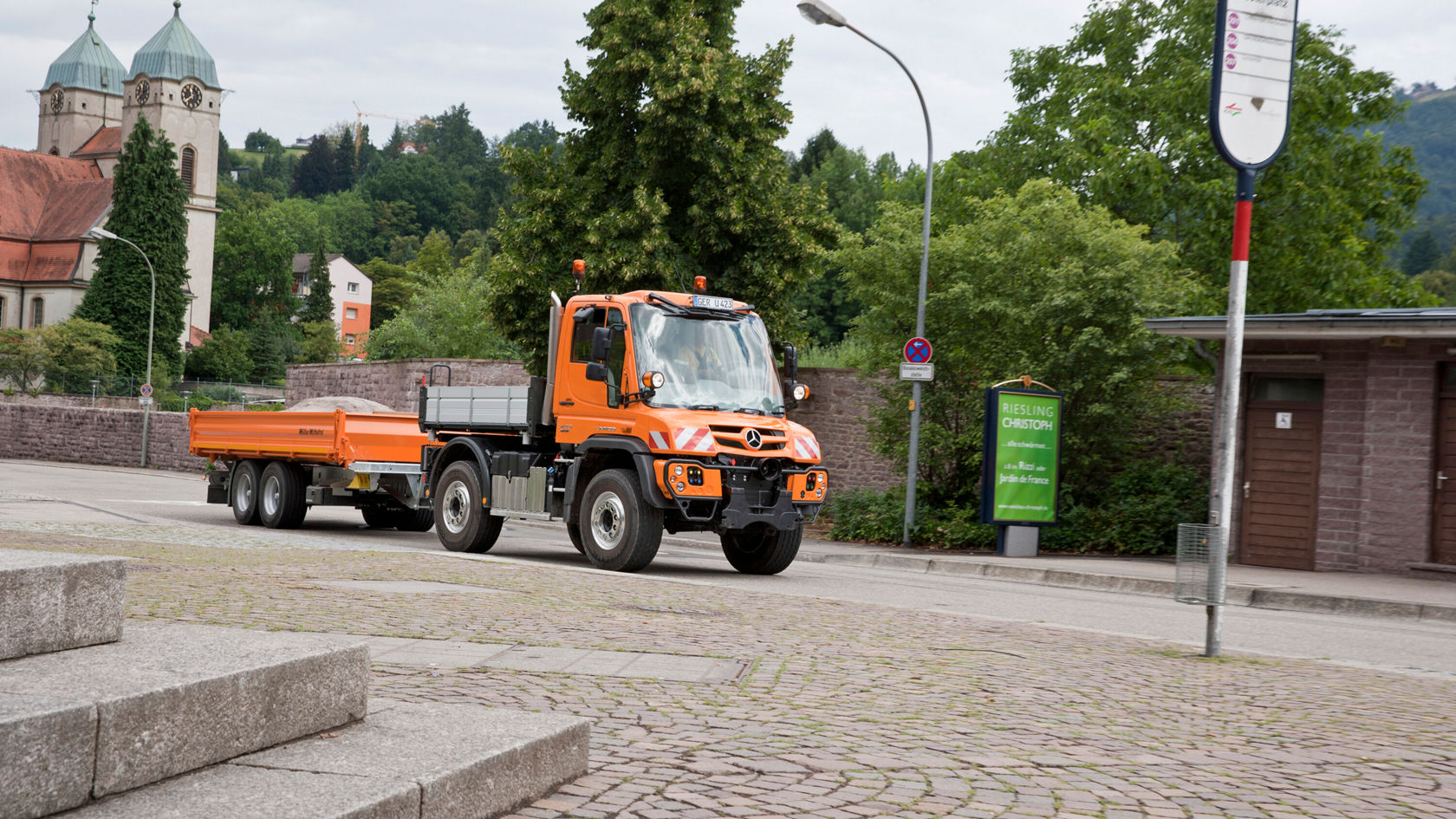 Un Unimog Mercedes‑Benz orange se trouve au bord d'une route, avec une remorque pour le transport de matériaux. Le Unimog est positionné en milieu urbain.