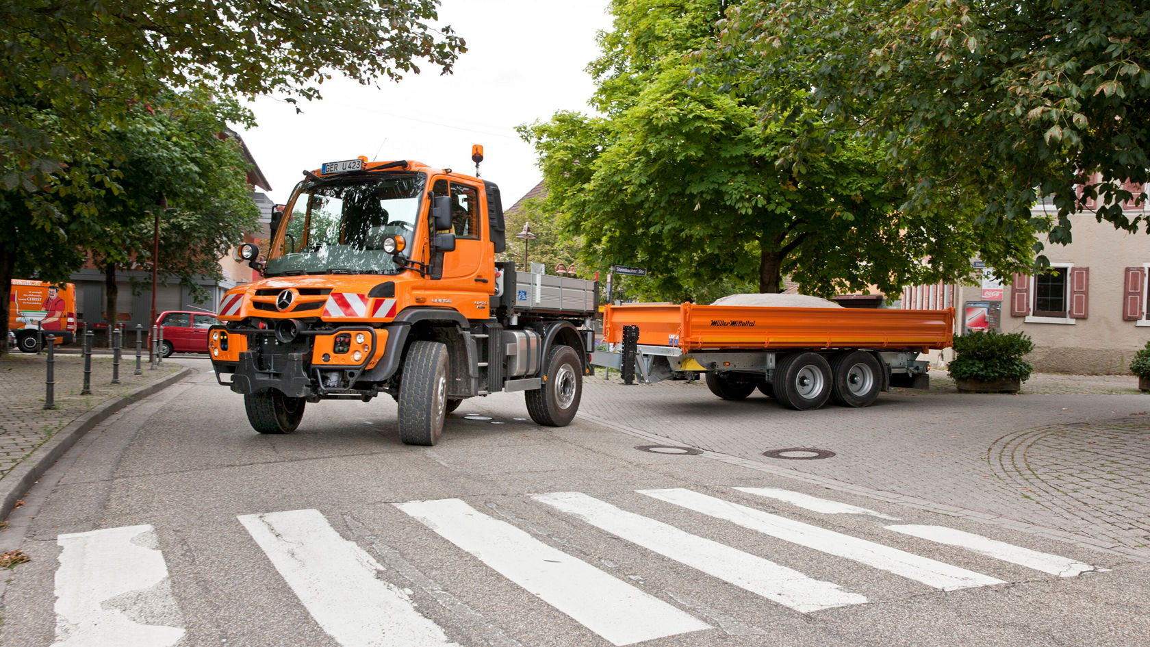 Un Unimog Mercedes‑Benz orange, avec une remorque, est arrêté à un carrefour, entouré d'arbres et d'immeubles d'habitation, et attend la circulation.