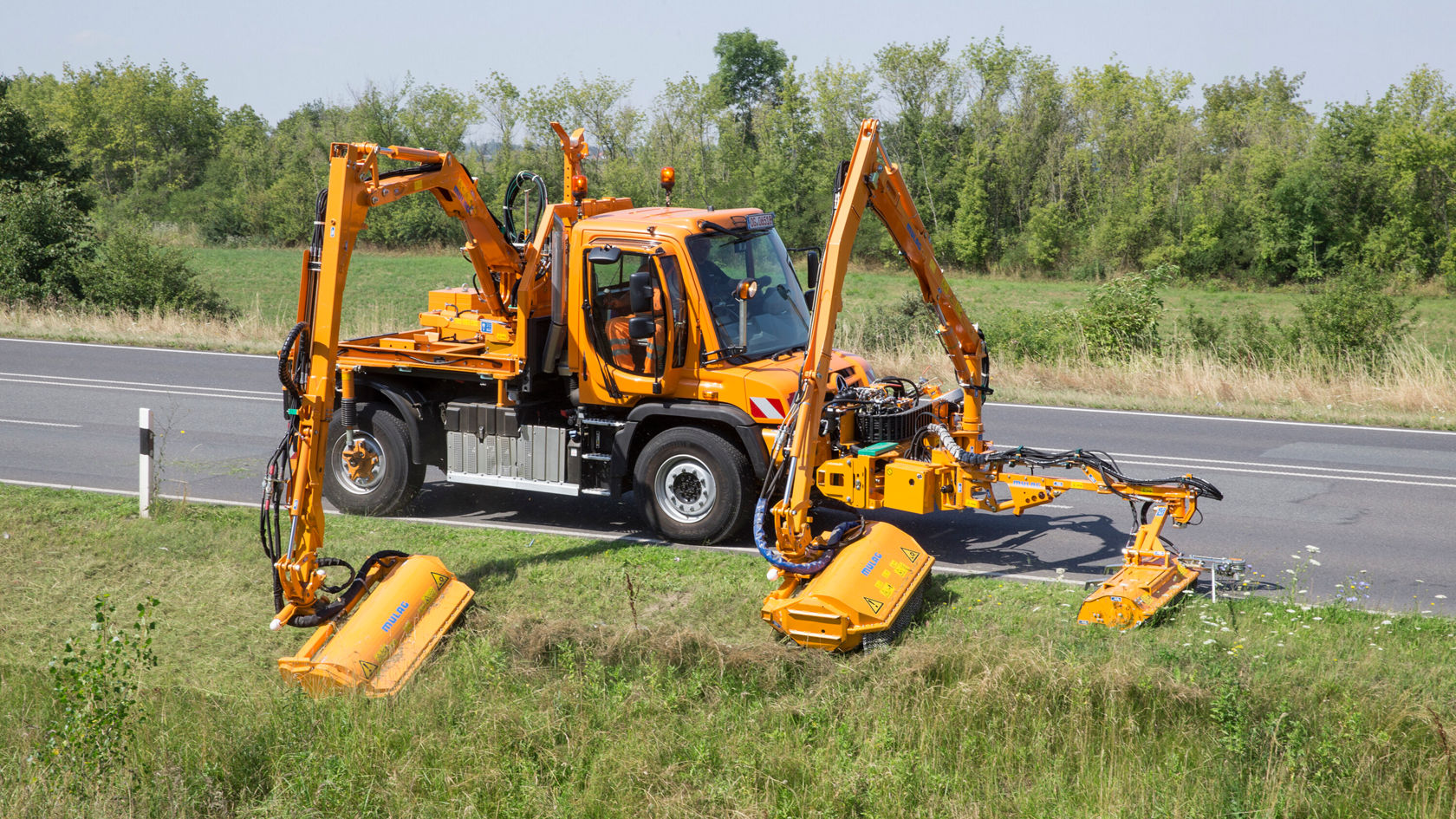 Un Unimog Mercedes‑Benz équipé de deux bras hydrauliques est stationné au bord de la route et effectue une opération de tonte et d'entretien, tout en coupant l'herbe le long de la chaussée.