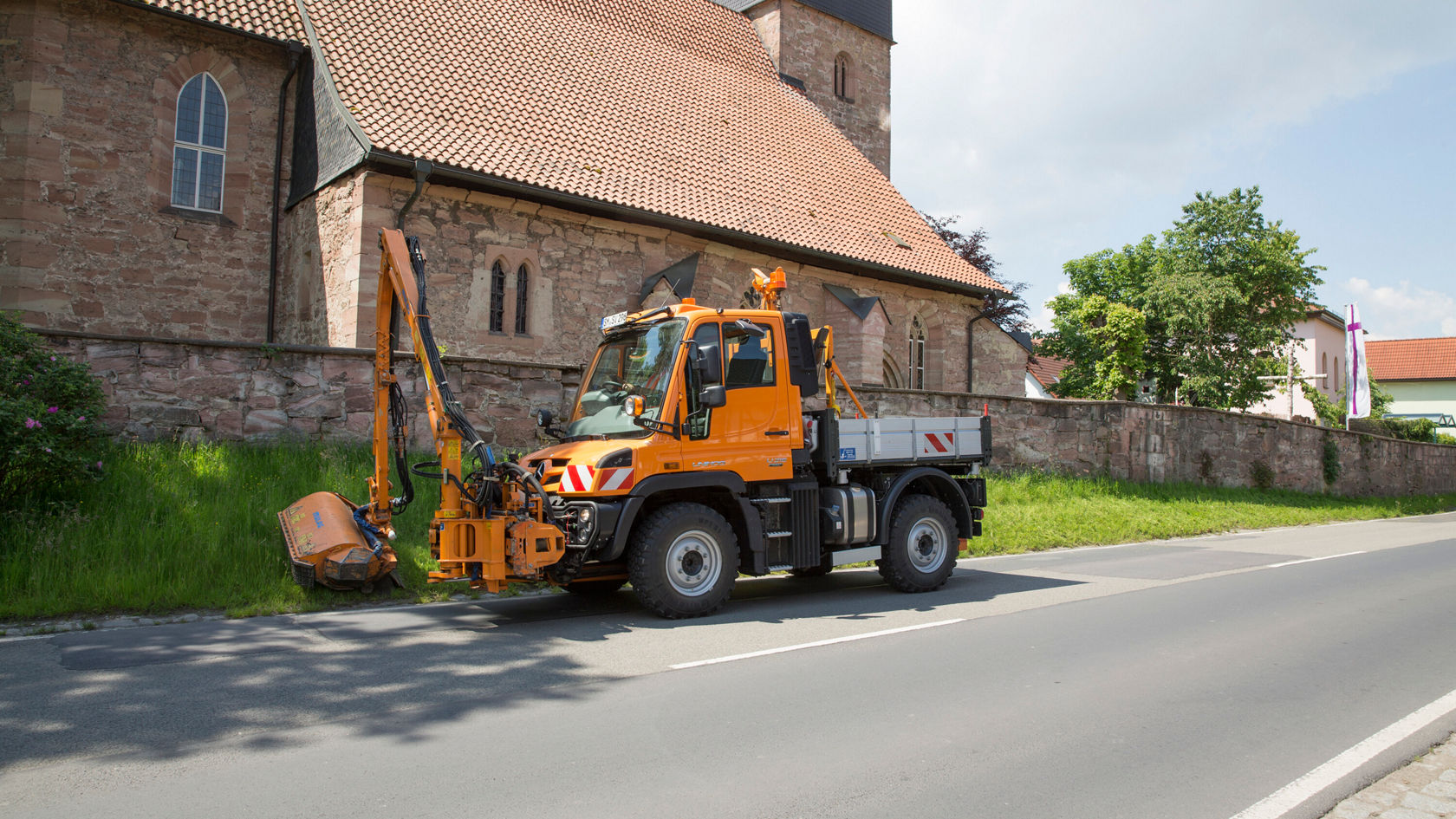 Un Unimog Mercedes‑Benz jaune est garé à côté d'une église, équipé d'un bras de travail et d'un outil rapporté pour le nettoyage du bord de la route.