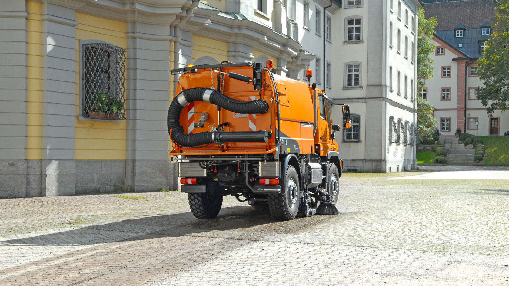 Un Unimog Mercedes‑Benz orange est stationné sur une rue pavée en ville. Il est équipé d'une grande installation d'évacuation d'air et se distingue par sa construction robuste.