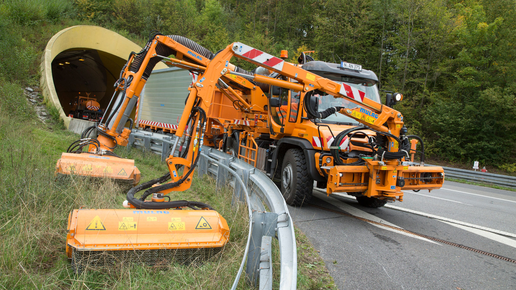 Un Unimog Mercedes‑Benz doté d'une superstructure orange est stationné au bord de la route, tandis que des bras mécaniques travaillent à l'ouverture d'un tunnel. Des panneaux d'avertissement de sécurité sont bien visibles.