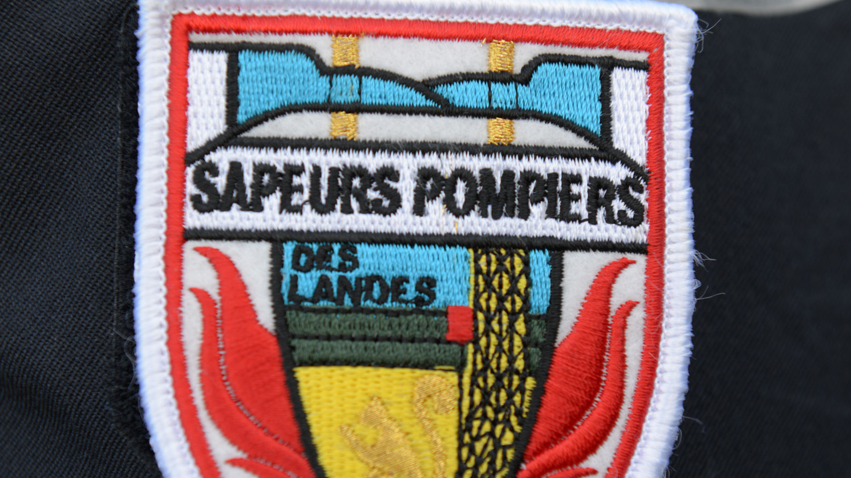 Abzeichen der Feuerwehr "Sapeurs-Pompiers des Landes" mit symbolischen Elementen wie Flammen und einer Grafik von Besatzungsfahrzeugen in den Farben Blau, Gelb und Rot.