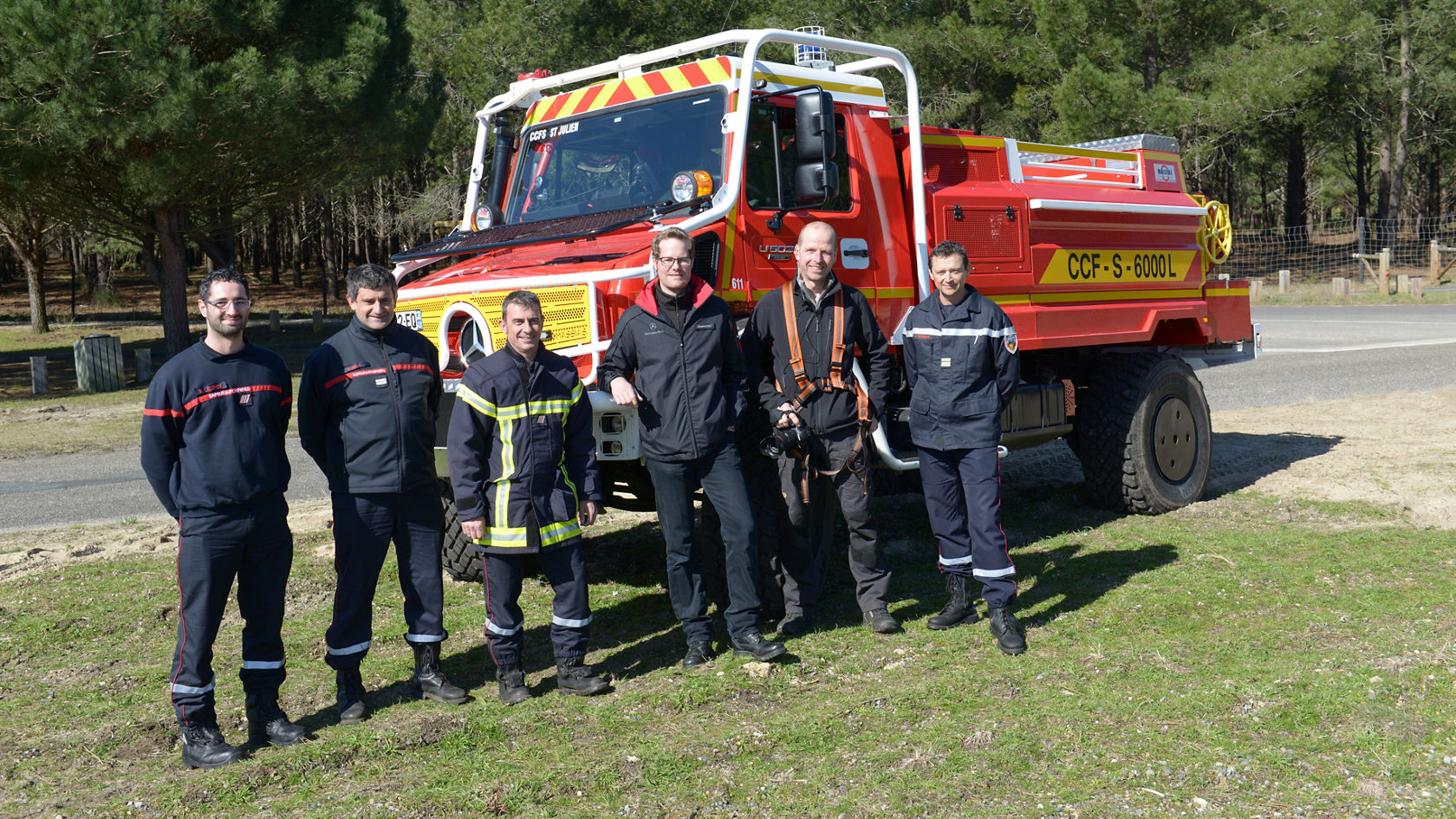 Sechs Feuerwehrleute in Uniform stehen neben einem roten Löschfahrzeug von Mercedes-Benz in einem Waldgebiet.