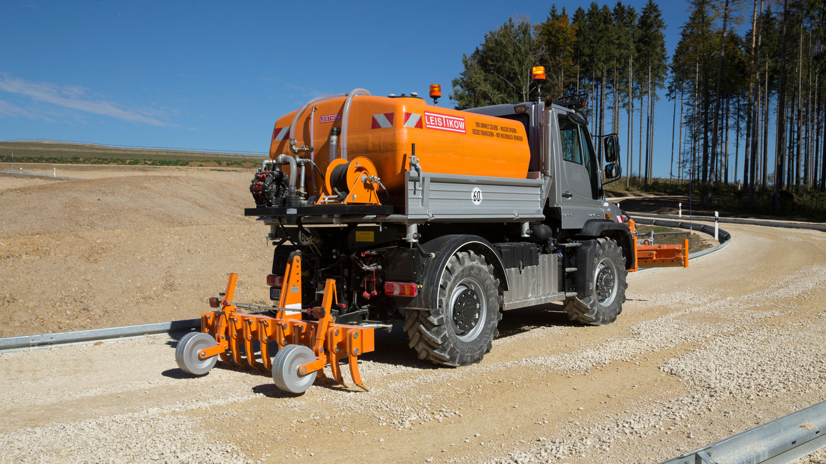 Un Unimog Mercedes‑Benz, équipé d'un réservoir orange et d'un dispositif de déblaiement, circule sur une route gravillonneuse dans un paysage forestier clairsemé.