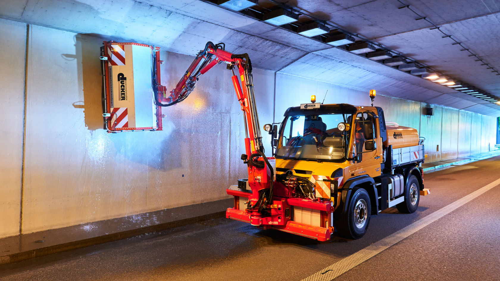 Ein Mercedes-Benz Unimog mit einem hydraulischen Arm reinigt die Wand eines Tunnels, während Scheinwerfer auf den Bereich gerichtet sind.