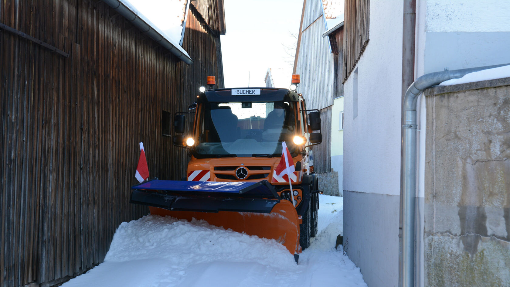 Un Unimog Mercedes‑Benz équipé d'une lame de déneigement dégage la neige d'une ruelle étroite. Des drapeaux rouges sont fixés sur les côtés du véhicule.