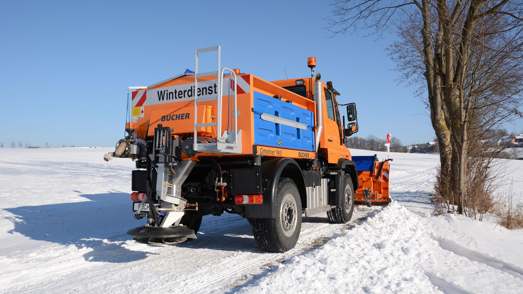 Un Unimog Mercedes‑Benz orange se trouve sur une route enneigée, équipé d'une lame biaise et d'un réservoir pour les produits de salage.