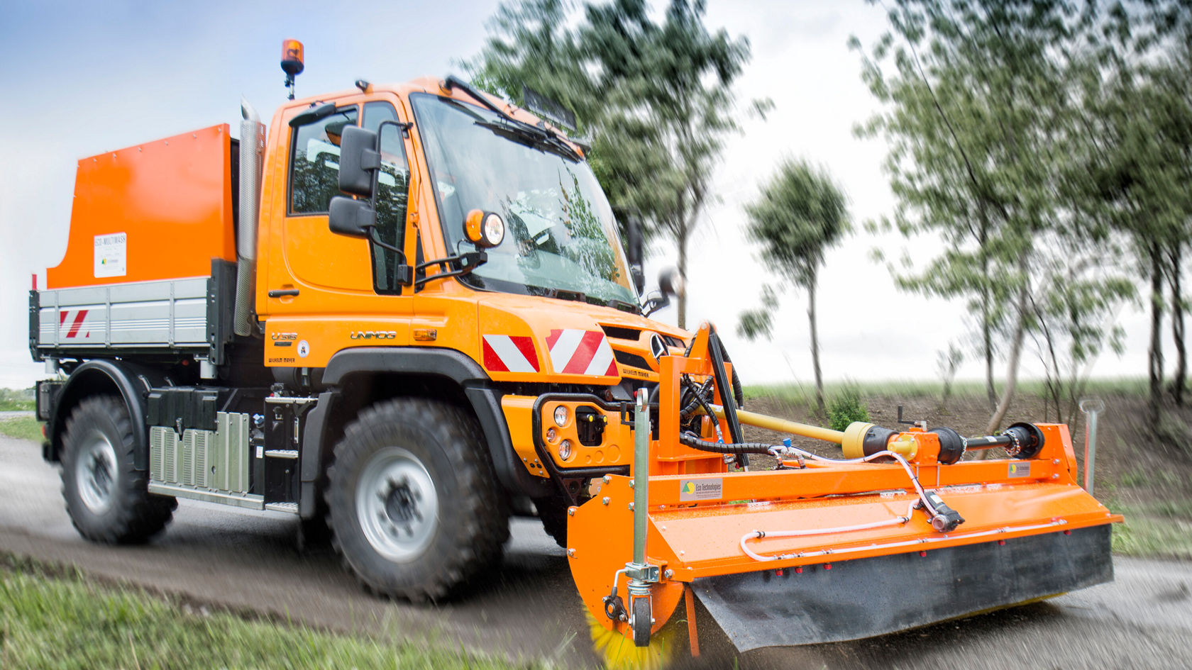 Ein Mercedes-Benz Unimog fährt auf einer Straße und ist mit einer orangefarbenen Kehrmaschine ausgestattet, die am Fahrzeug angebracht ist und den Boden bearbeitet.