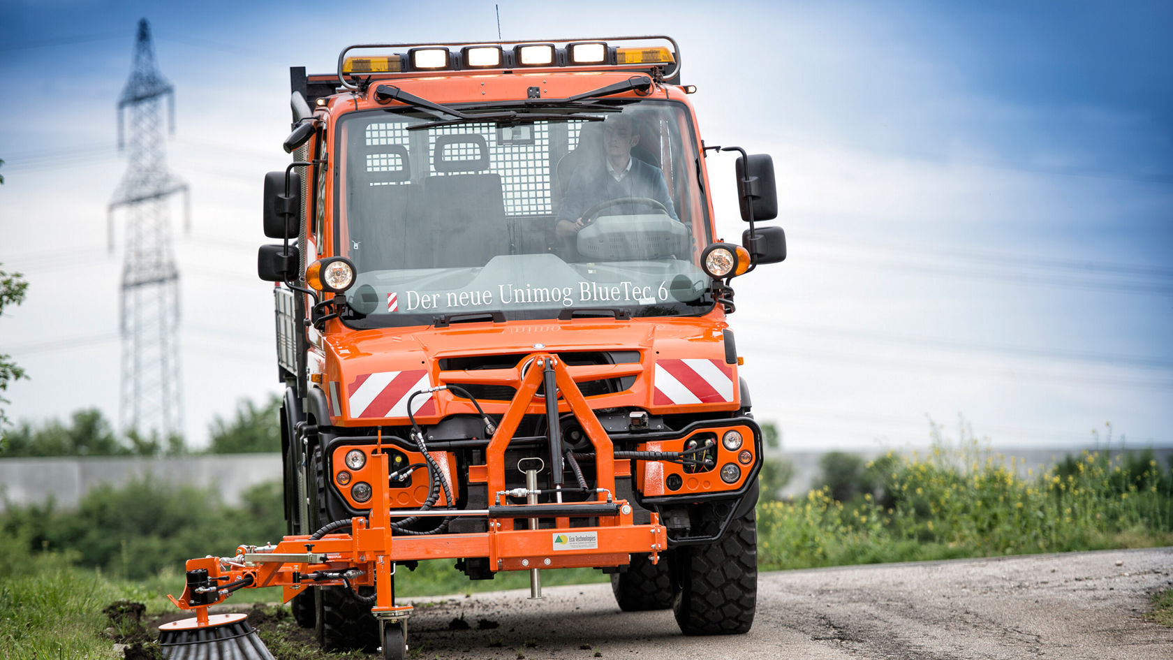 Un Unimog Mercedes-Benz effectue un nettoyage de la chaussée, une brosse enlevant la saleté du bord de la route.