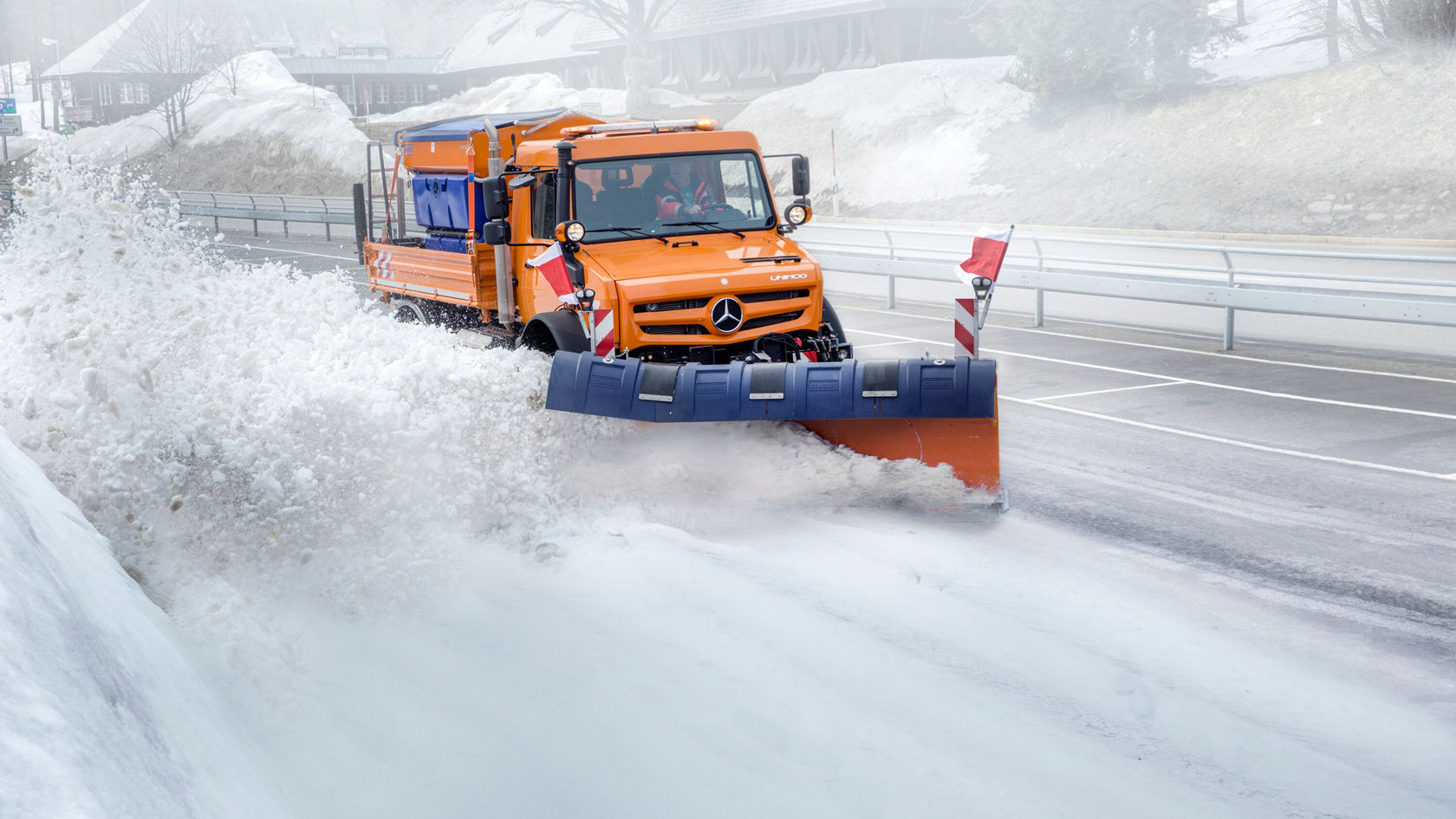 Un Unimog Mercedes‑Benz dégage une piste enneigée à l'aide d'une lame de déneigement, tandis que la neige est repoussée de chaque côté de la piste.