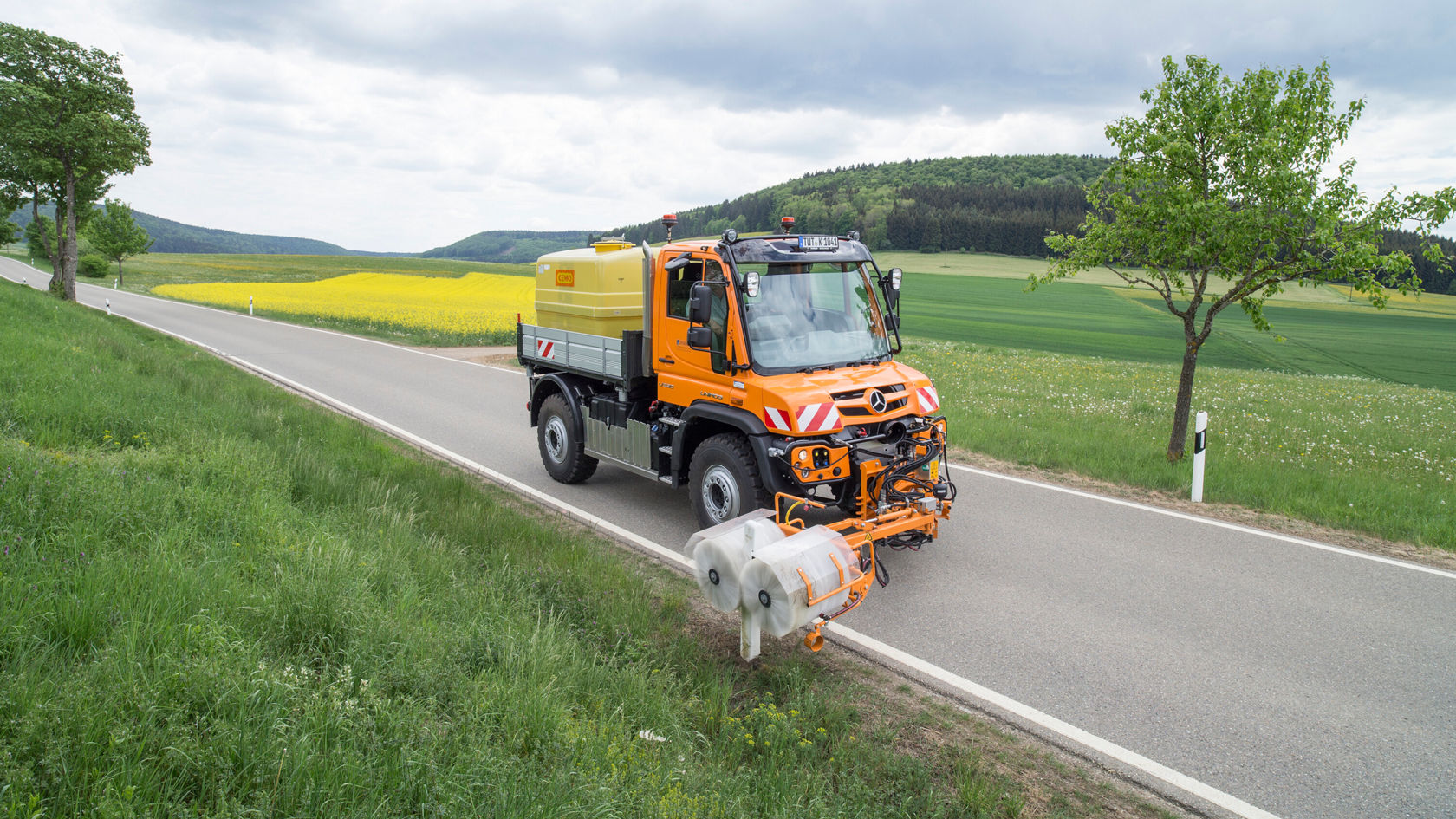 Ein orangefarbener Mercedes-Benz Unimog steht auf einer Landstraße. Der Unimog ist mit einem Wassertank und einer Arbeitsvorrichtung ausgestattet, um Straßen zu reinigen.