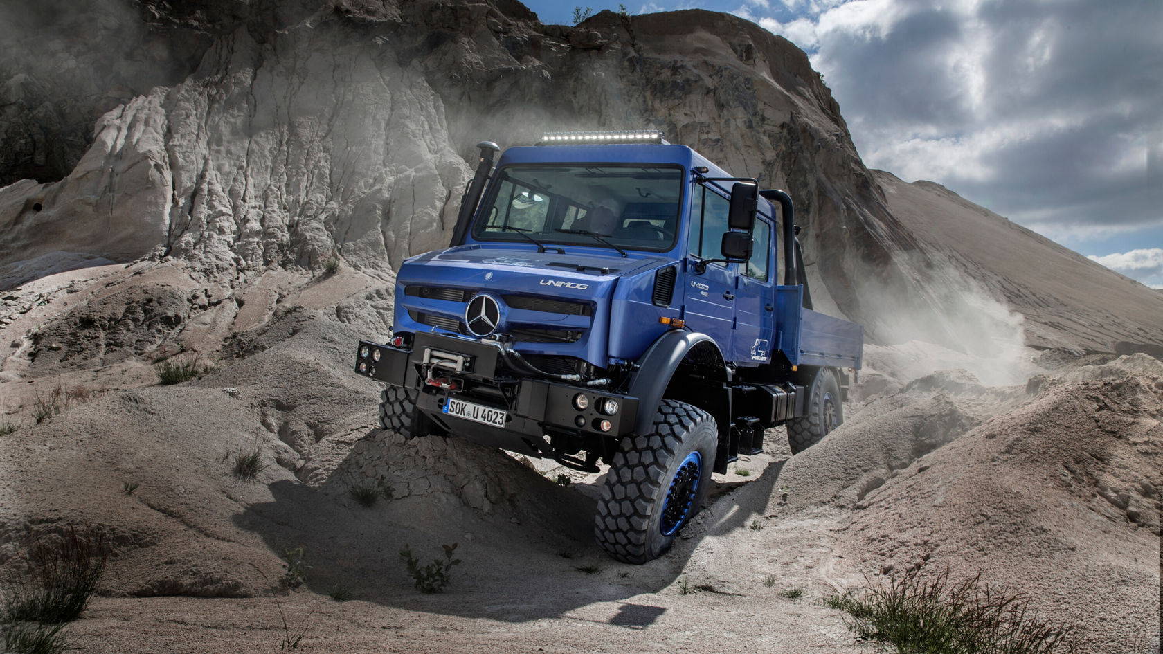 Ein blauer Unimog U 4023 mit großen Reifen fährt durch sandiges Terrain mit felsigen Hintergründen.
