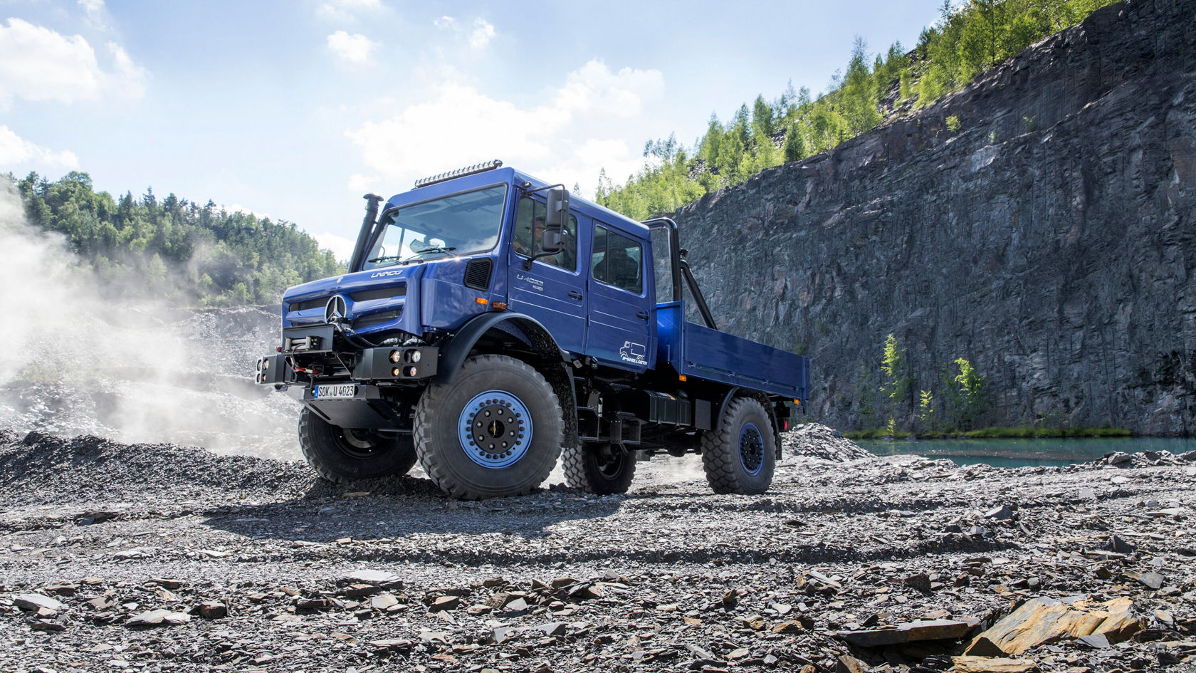 Ein blauer Unimog U 4023 steht auf einem kiesbedeckten Gelände, umgeben von einer steilen Felswand und Bäumen im Hintergrund. 