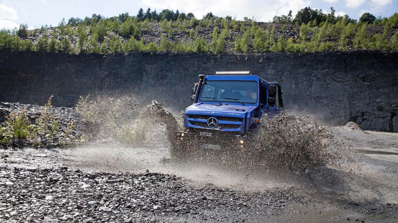 Ein blauer Unimog U 4023 fährt durch schlammiges Terrain, wobei er Spritzer und Schmutz aufwirbelt. 