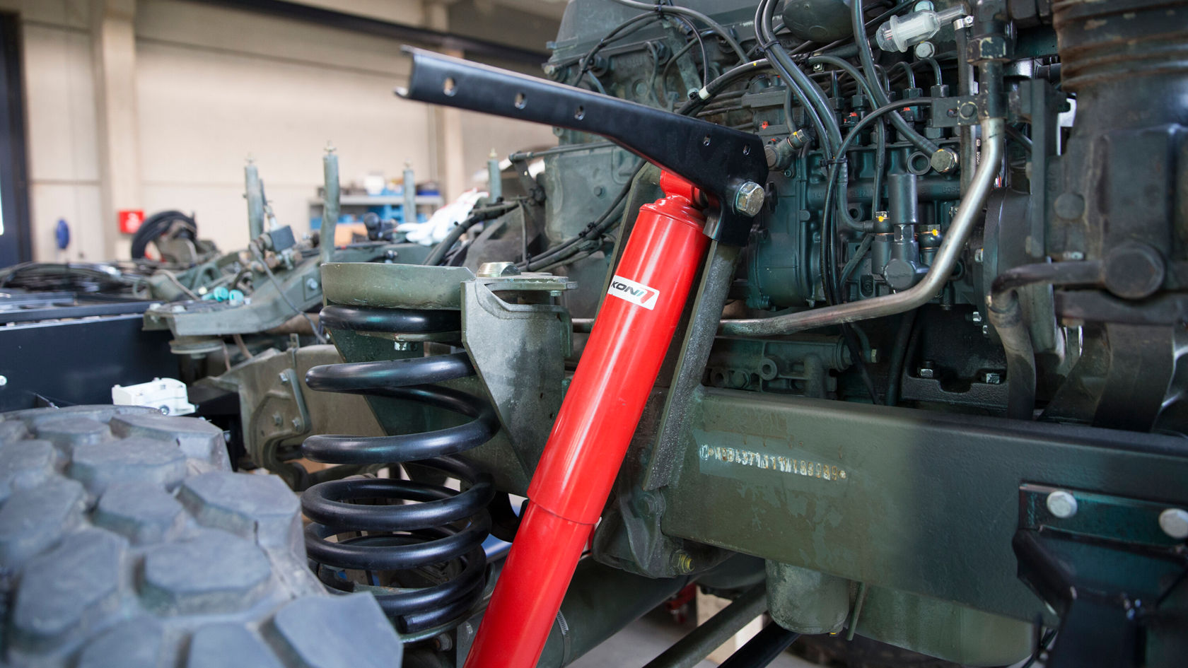 Ein Fahrzeugmotors in einer Werkstatt. Im Vordergrund ist ein roter Stoßdämpfer sichtbar, der an einer Feder und mehreren Motorkomponenten befestigt ist. Im Hintergrund sind Werkstattutensilien und weitere Fahrzeugteile zu erkennen.