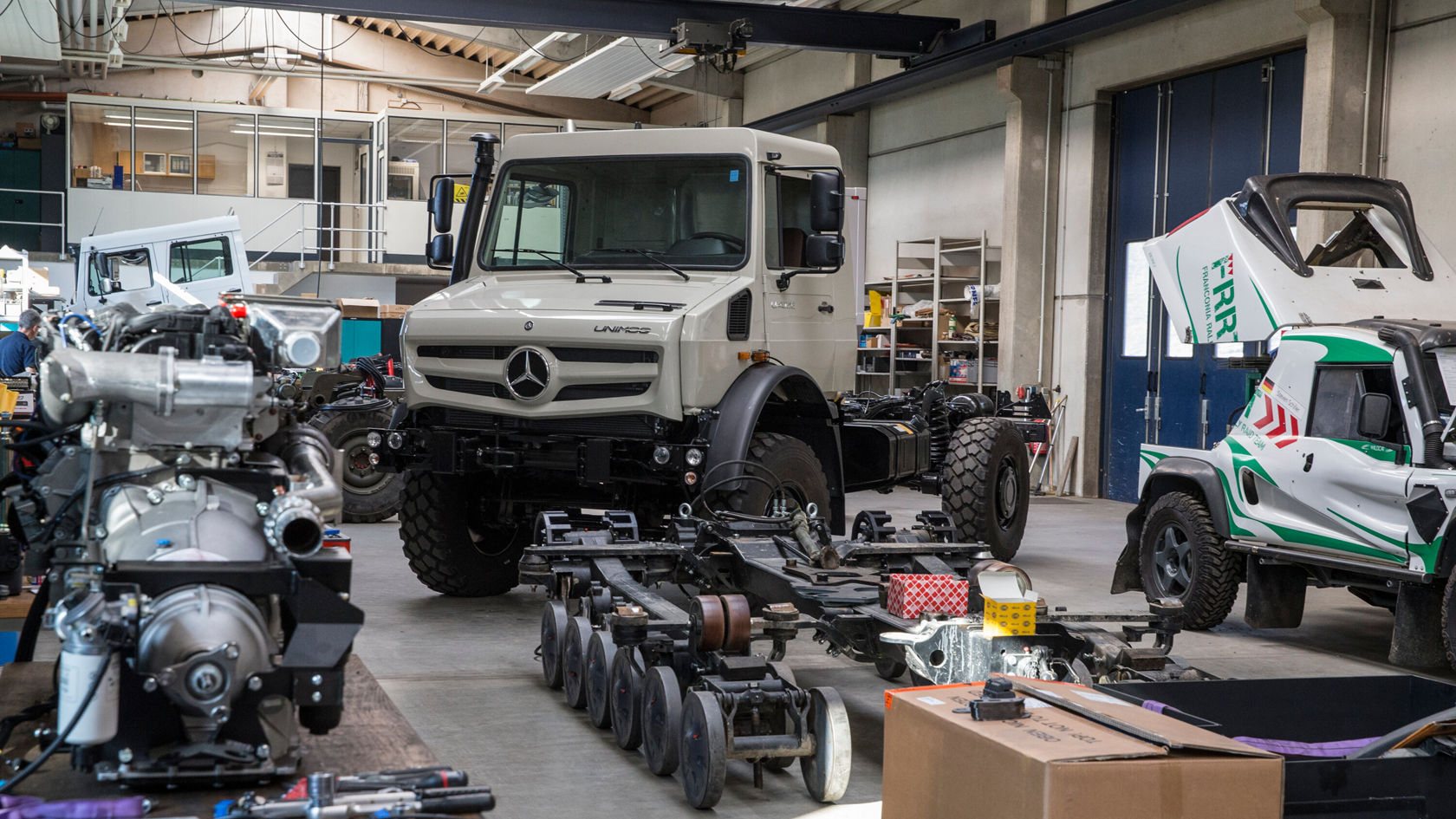 In einer Werkstatt stehen verschiedene Fahrzeuge und Teile. Im Vordergrund ist ein großer Mercedes-Benz Unimog zu sehen, umgeben von einem Motor und einem mechanischen Gerät.