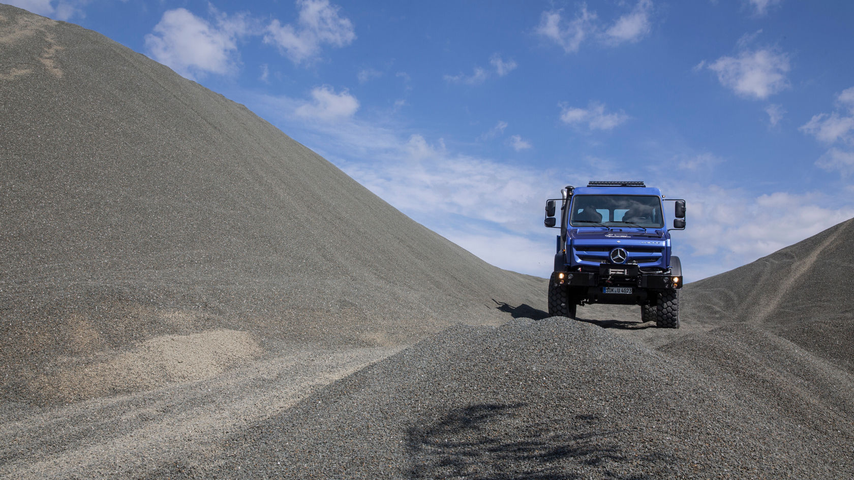 Ein blauer Unimog U 4023 steht auf einem kleinen Hügel aus Schotter, umgeben von größeren Schotterhaufen.