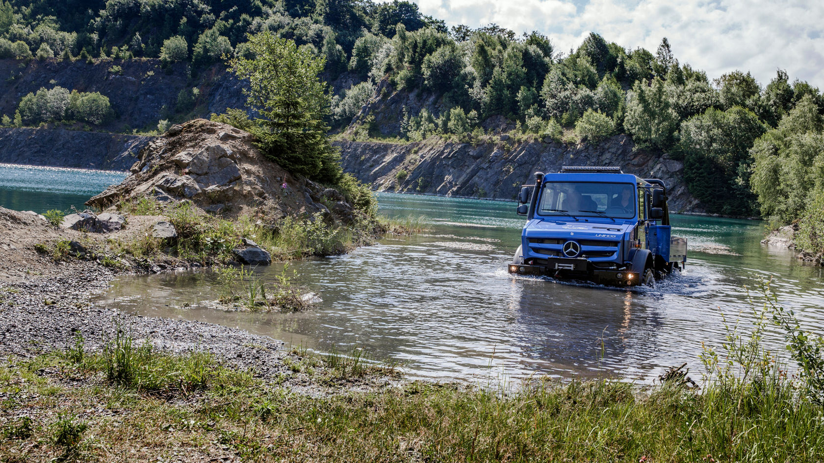 Ein blauer Unimog U 4023 fährt durch flaches Wasser in einem kleinen See. 