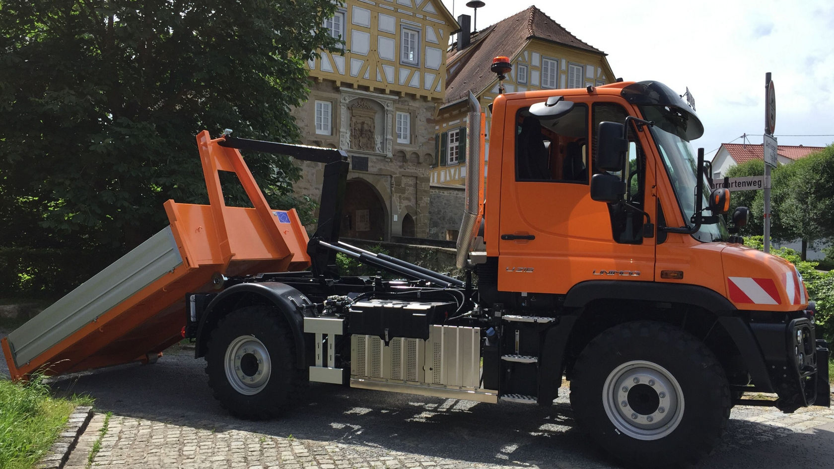 Ein orangefarbener Mercedes-Benz Unimog mit einer Kippmulde steht auf einer Straße. Die Kippmulde ist angehoben, um den Ladevorgang zu zeigen.