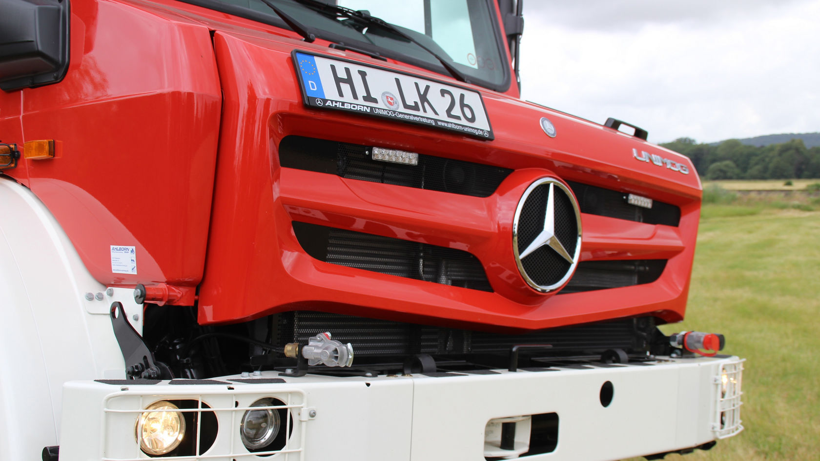 Roter Mercedes-Benz Unimog mit markantem Kühlergrill und Scheinwerfern, präsentiert in einer ländlichen Umgebung. Der Fokus liegt auf der Frontansicht des Fahrzeugs.
