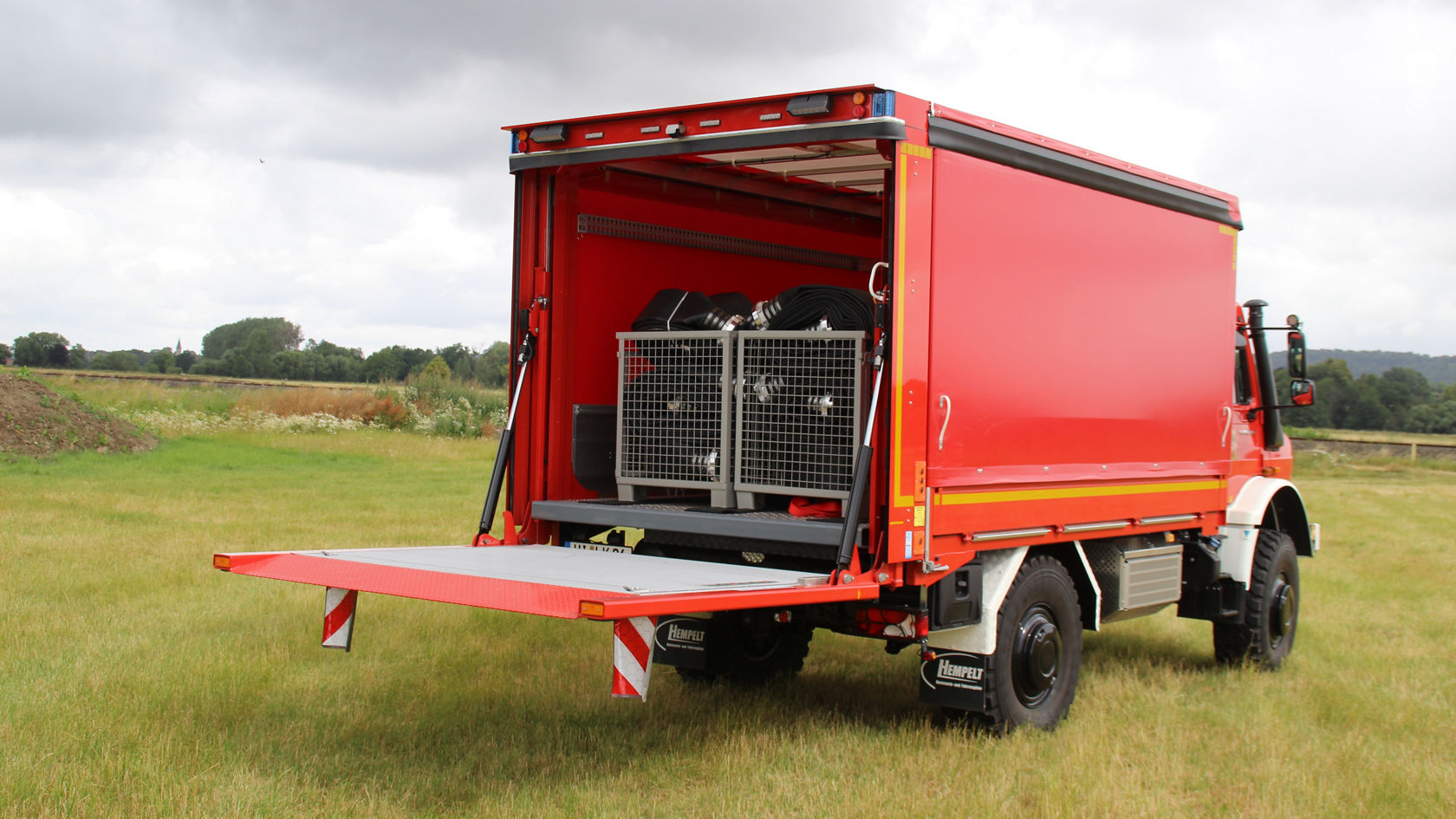 Ein roter Mercedes-Benz Lkw mit geöffneter Ladefläche steht auf einer Wiese. Die Ladefläche zeigt einige Käfige. Der Lkw ist für den Transport optimiert.