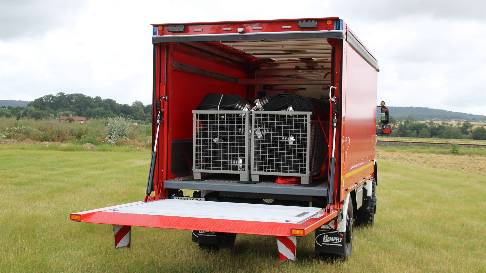 Der geöffneten hintere Bereich eines roten Mercedes-Benz Lkw zeigt zwei Gitterboxen mit Schläuchen. Eine ausgeklappte Ladefläche ermöglicht den schnellen Zugang zu den Geräten.