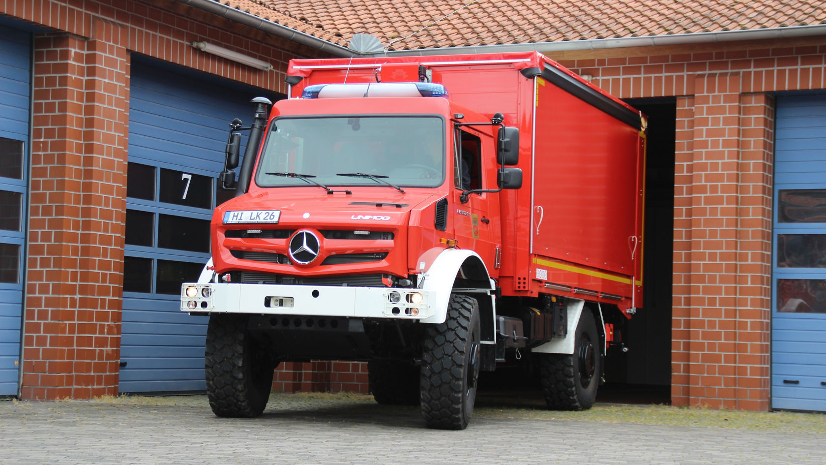 Roter Mercedes-Benz Einsatzfahrzeug steht vor einer Feuerwache, teilweise in der Garage. Das Fahrzeug hat große Reifen und eine Einsatzbeleuchtung auf dem Dach.