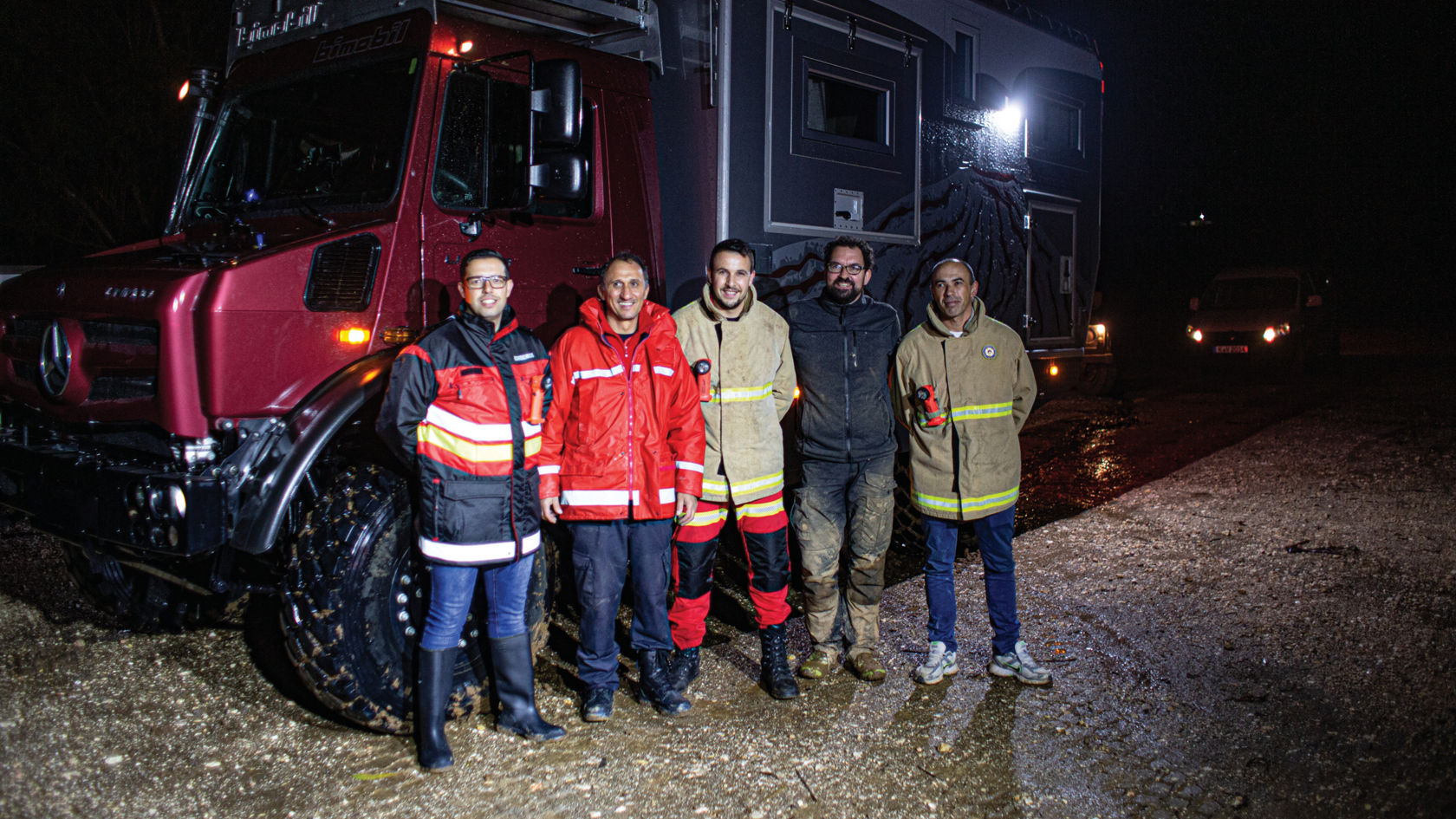 Een groep van vijf personen staat glimlachend voor een Mercedes-Benz voertuig in de nacht, terwijl op de achtergrond een mobilhome te zien is. Ze dragen inzetkledij.