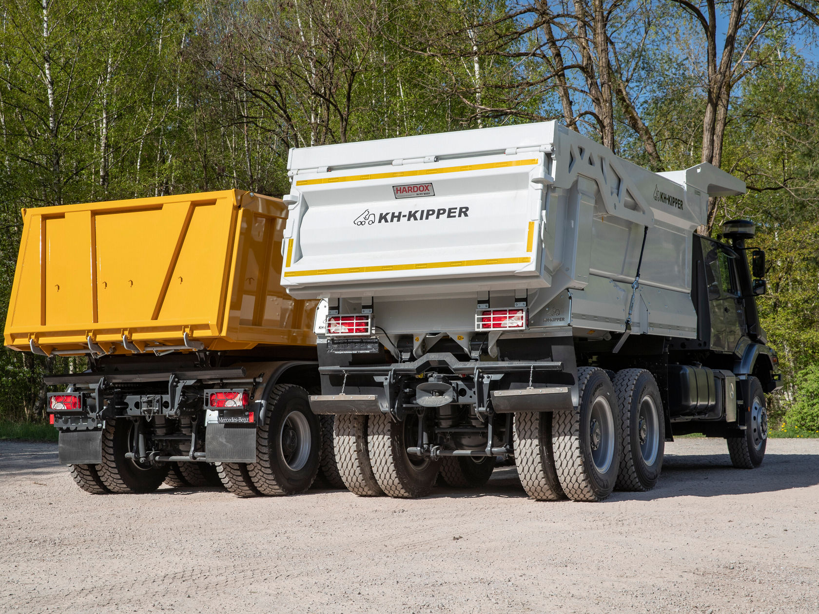 Zwei große Lkw mit Kippmulden, einer gelb und einer weiß, stehen nebeneinander auf einem Kiesplatz, umgeben von Bäumen. Die Fahrzeuge sind für den Transport von Schüttgütern ausgelegt.