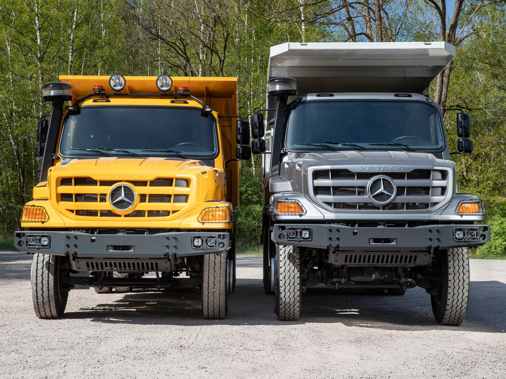 Zwei Mercedes-Benz Nutzfahrzeuge stehen nebeneinander, ein gelbes und ein silbernes Modell, beide mit robustem Design und großen Kühlergrills, auf einer asphaltierten Fläche umgeben von Bäumen.