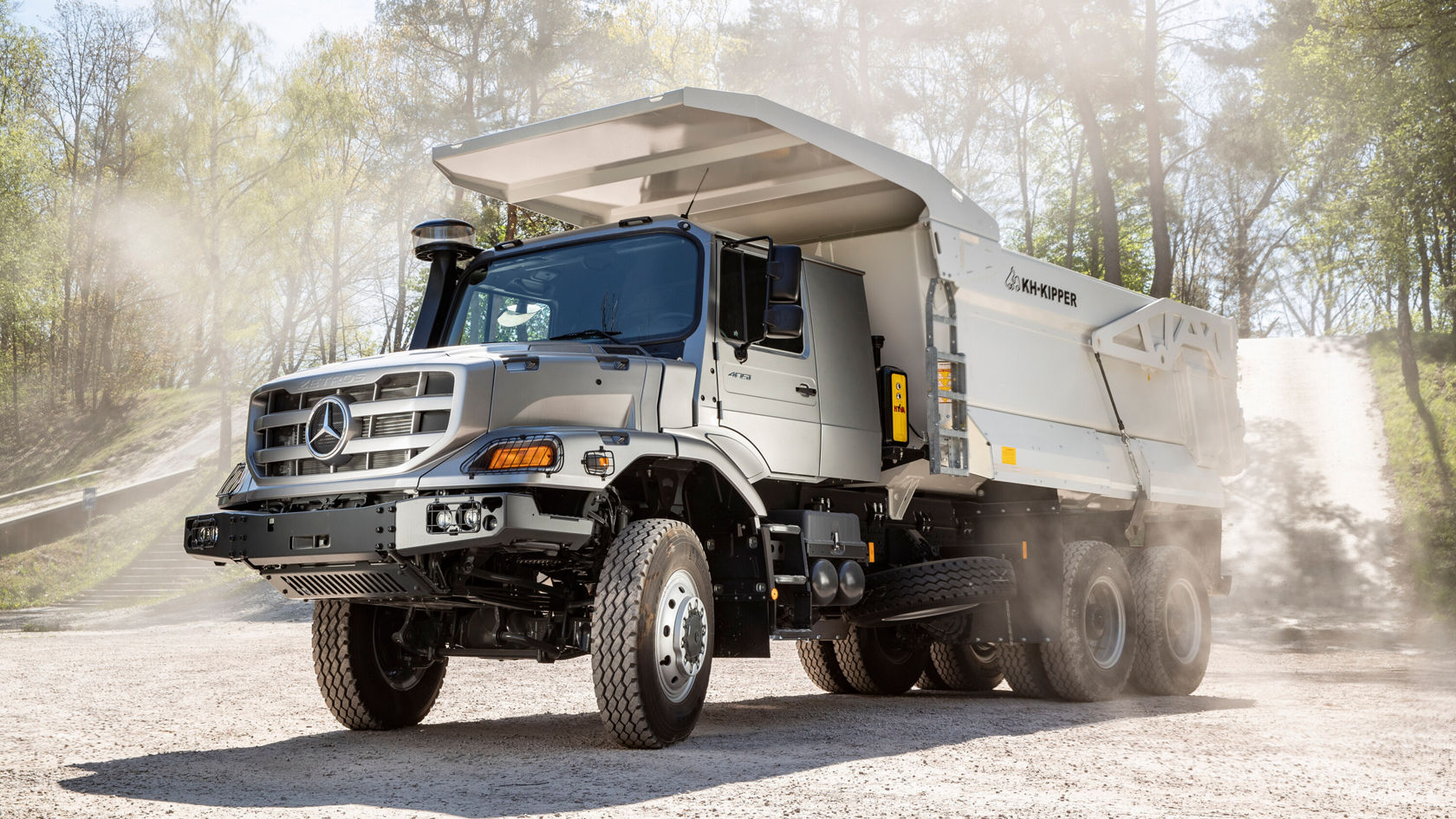 Ein großer Mercedes-Benz Lkw mit Kipperaufbau steht auf einer staubigen Fläche. Der Kipper ist angehoben und es sind Bäume im Hintergrund sichtbar.