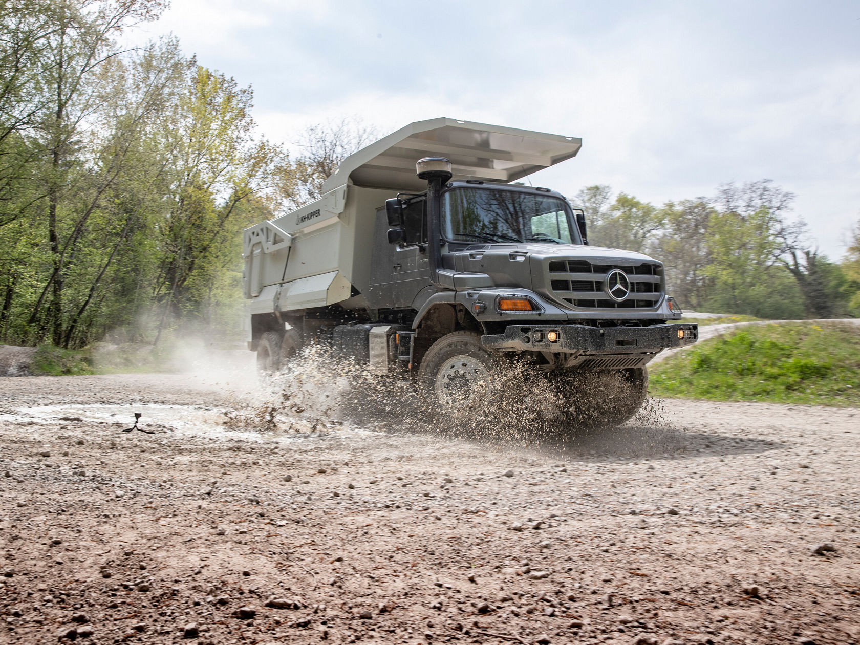 Ein Mercedes-Benz Lkw fährt durch schlammigen Untergrund und spritzt Wasser und Schlamm auf, umgeben von Bäumen und einer natürlichen Landschaft.