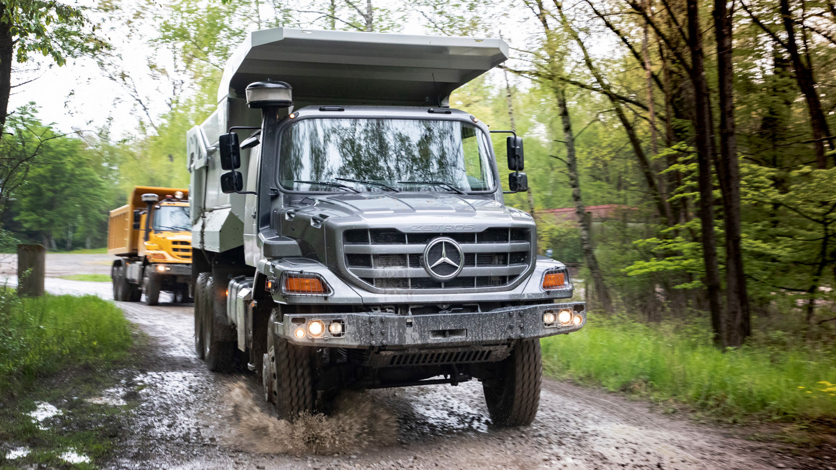 Ein Mercedes-Benz Kipper fährt durch eine schlammige, unbefestigte Straße, während ein weiterer Lkw im Hintergrund zu sehen ist. Die Umgebung ist von Bäumen und Grün geprägt.