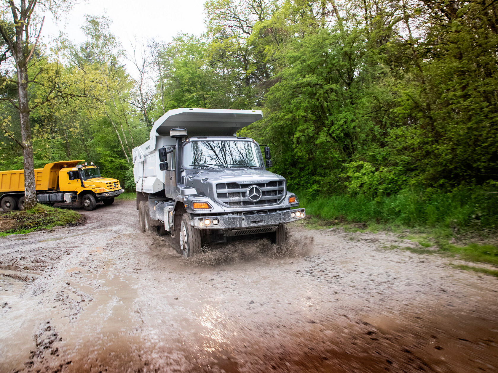 Ein Mercedes-Benz Kipper fährt durch einen schlammigen Weg in einem bewaldeten Gebiet, während ein gelber Lkw im Hintergrund sichtbar ist.