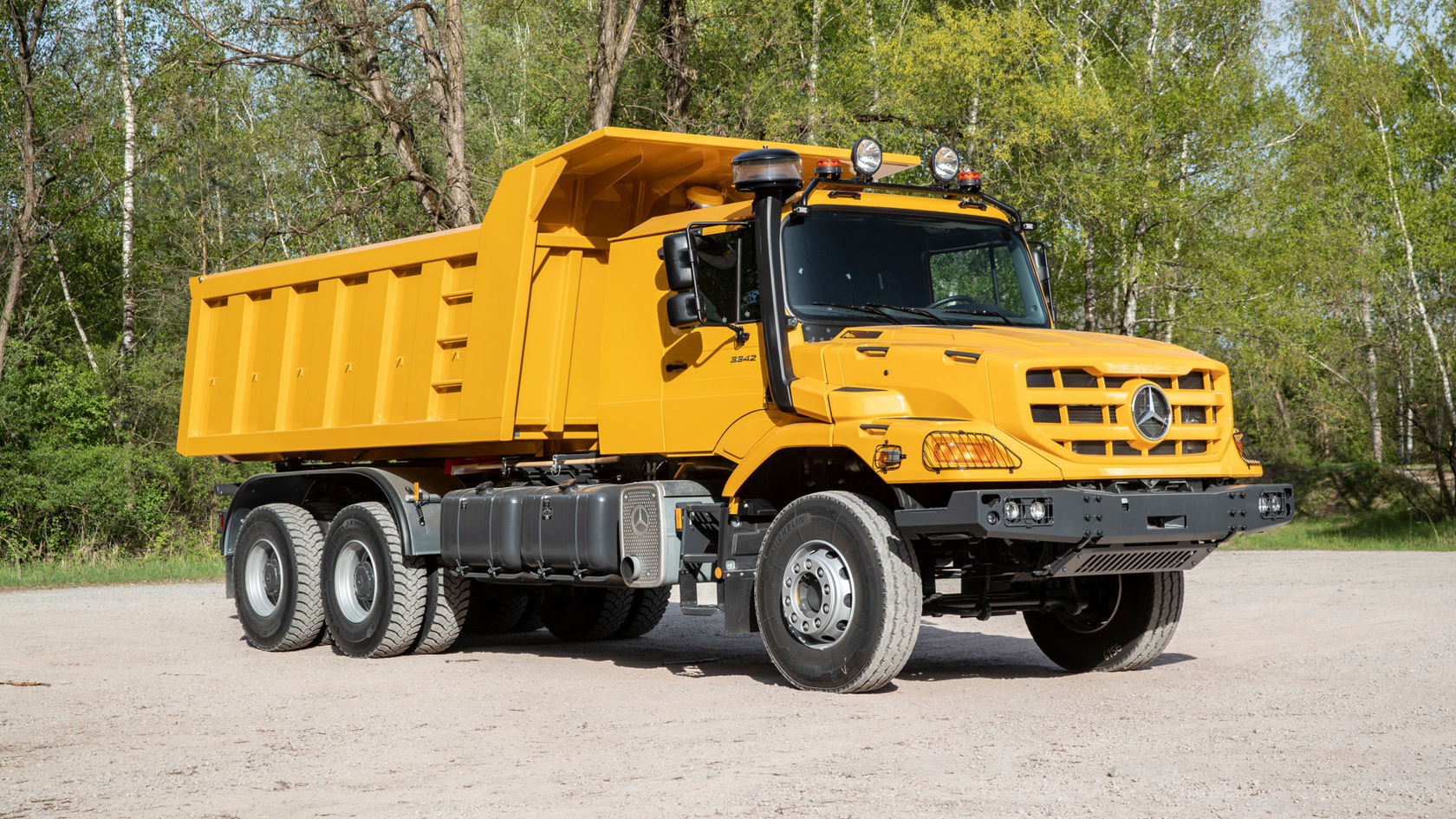 Ein gelber Mercedes-Benz Kipper steht auf einer unbefestigten Fläche, umgeben von Bäumen. Der Lkw hat eine Kabine mit mehreren Scheinwerfern und einer robusten Bauweise.
