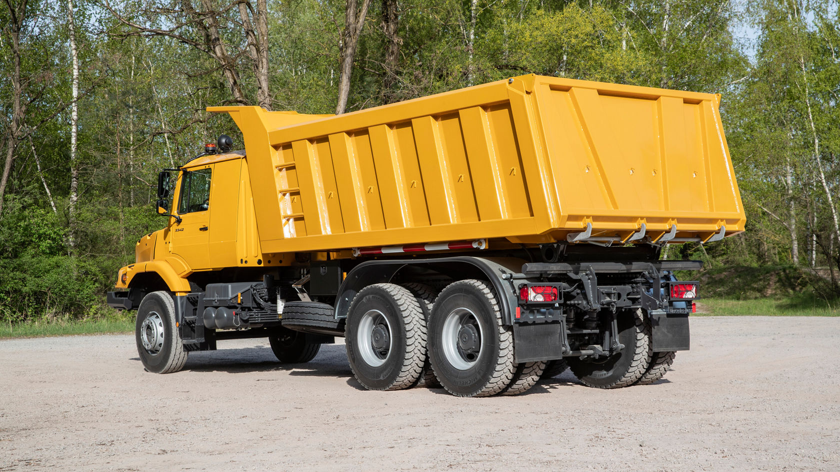 Ein gelber Kipplaster steht auf einer unbefestigten Fläche, umgeben von Bäumen. Der Laster hat eine breite, offene Ladefläche und ist bereit für den Transport von Materialien.