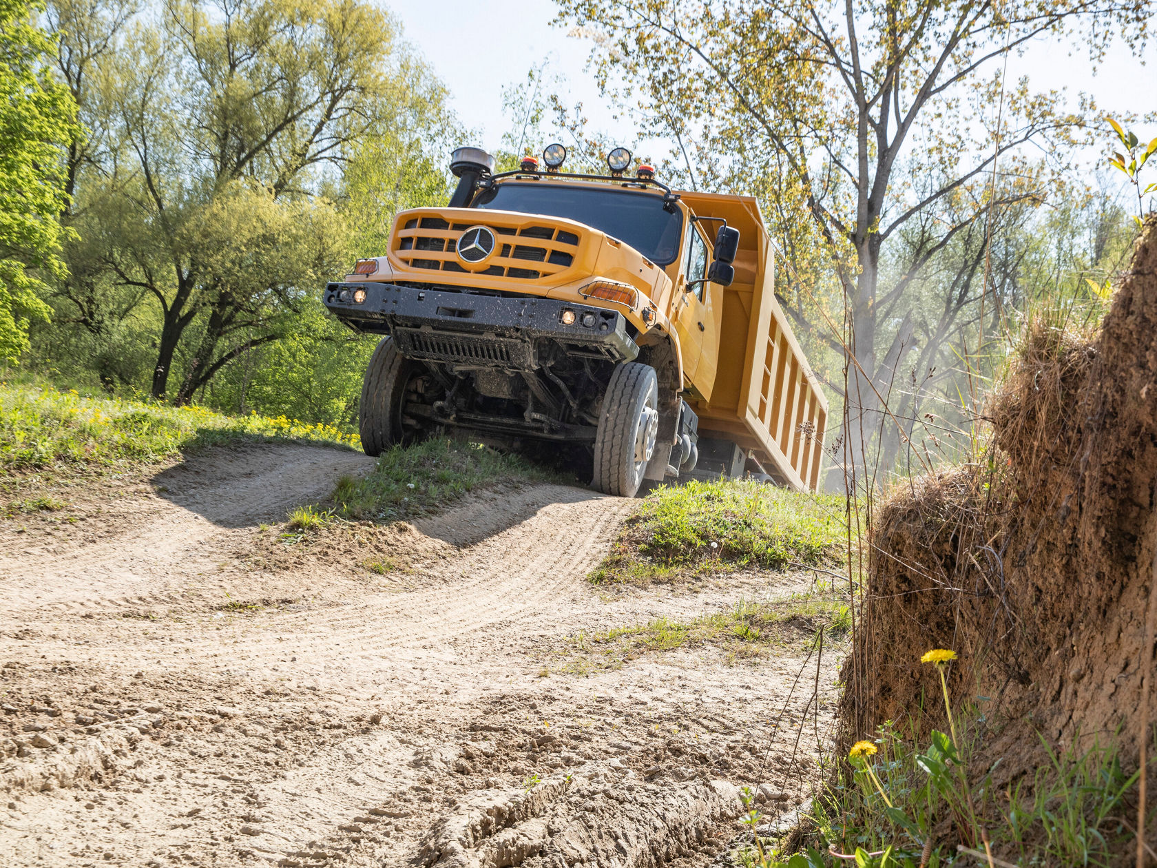 Ein gelber Mercedes-Benz Lkw fährt eine schottrige Piste entlang, umgeben von Bäumen und strahlendem Sonnenlicht, während er eine steile Neigung hinauffährt.
