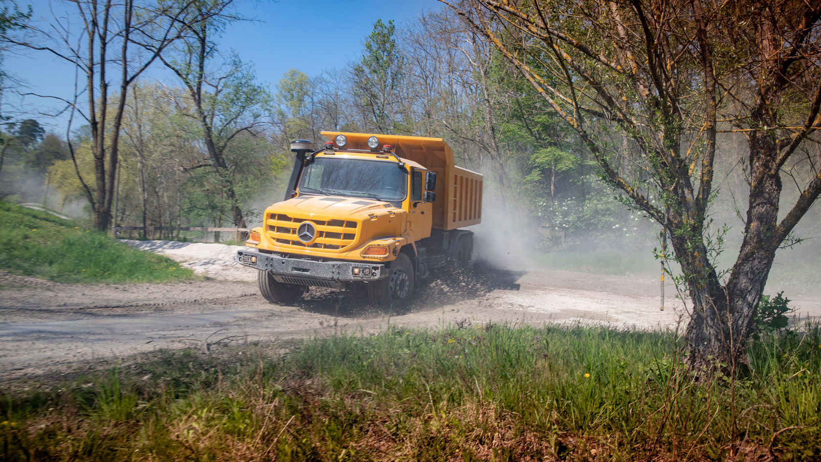Ein gelber Mercedes-Benz Lkw fährt auf einer staubigen Straße, während er eine Kurve macht und Staub aufwirbelt. Der Lkw ist für den Einsatz im Gelände geeignet.