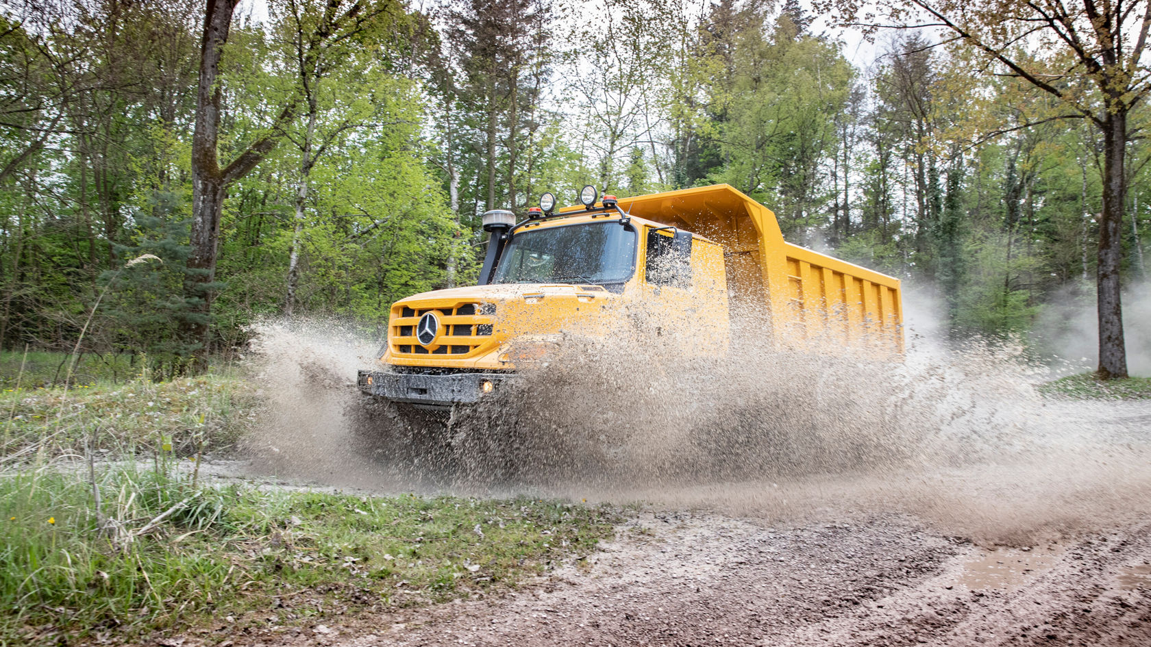 Ein gelber Mercedes-Benz Lkw fährt durch eine schlammige Pfütze im Wald und spritzt Wasser in die Luft, während er auf einem unbefestigten Weg unterwegs ist.