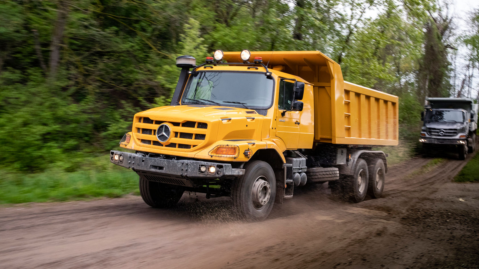 Ein gelber Lkw von Mercedes-Benz fährt auf einem unbefestigten Weg, umgeben von Bäumen. Der Lkw hat eine offene Ladeklappe und wirbelt Staub auf.