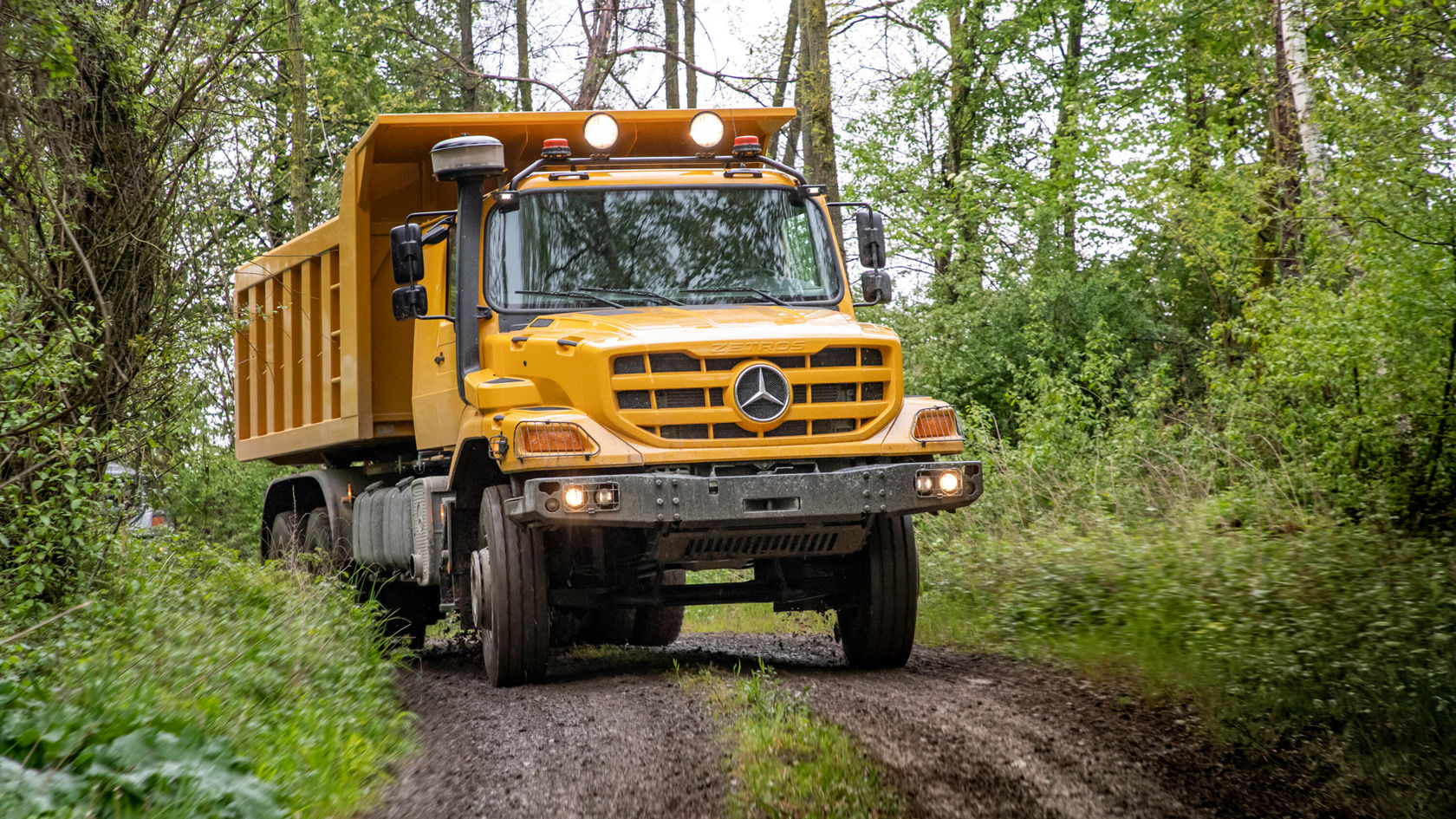 Ein gelber Mercedes-Benz Kipper fährt über einen schmalen, unbefestigten Weg, umgeben von dichter Vegetation. Der Lastwagen zeigt starke Geländetauglichkeit und hat eine robuste Bauweise.