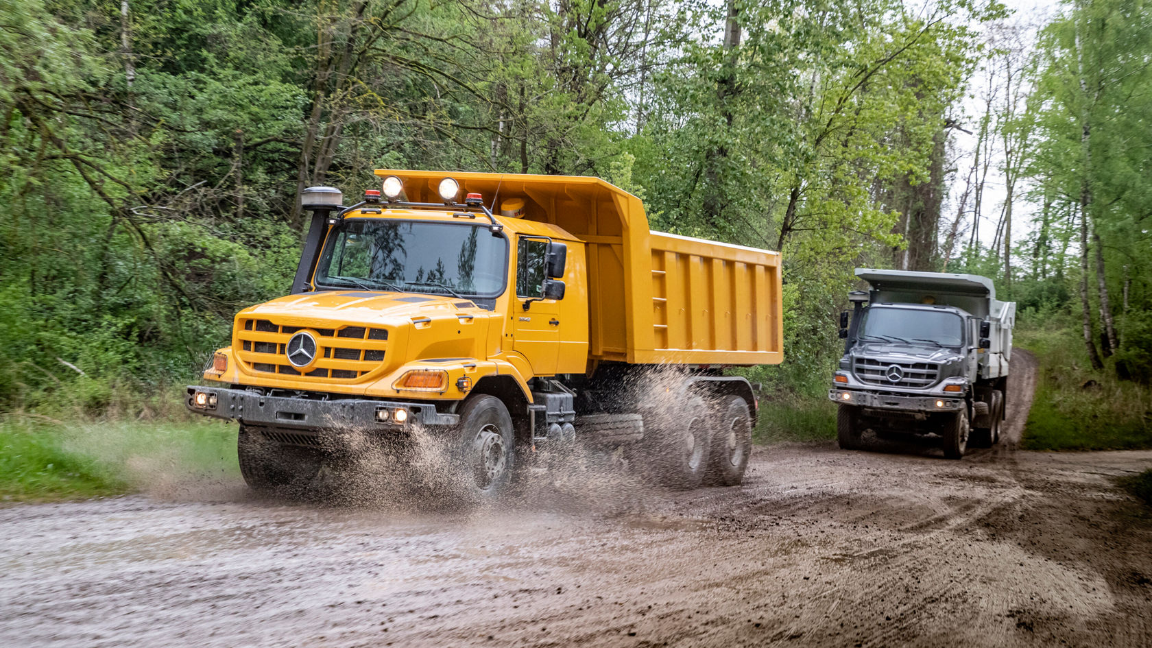 Ein gelber Mercedes-Benz Kipplaster fährt auf einem schottrigen Weg und spritzt dabei Schlamm, während ein anderer Mercedes-Benz Lkw im Hintergrund zu sehen ist.