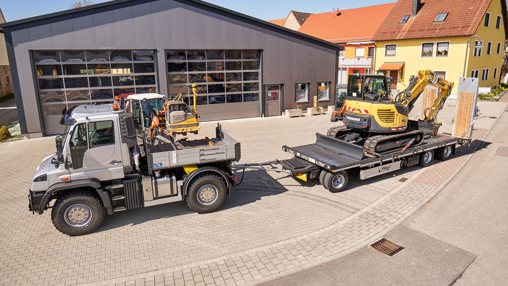 Un Unimog Mercedes‑Benz tire une remorque avec une pelleteuse sur une surface asphaltée devant un atelier, entouré de bâtiments d'habitation.