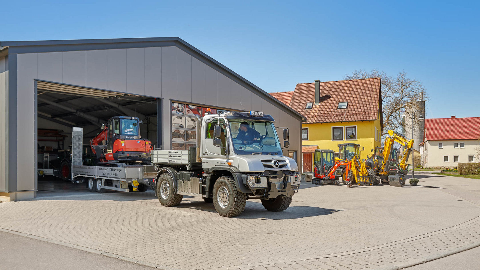 Un Unimog Mercedes-Benz circule devant un hall moderne en plein air, tractant une remorque sur laquelle est transportée une machine rouge, entouré de différents engins de terrassement.
