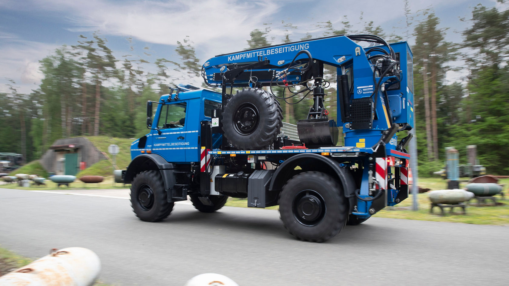 Un camión Mercedes-Benz azul con un implemento especializado para la desactivación de artefactos explosivos circula por una carretera, rodeado de árboles y objetos militares.