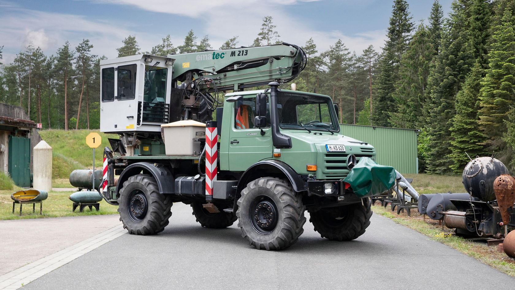Un Mercedes-Benz Unimog con un brazo de grúa está en una calle en un entorno industrial, rodeado de árboles y de varios equipos. El Unimog tiene una pintura verde y una cabina de mando blanca.