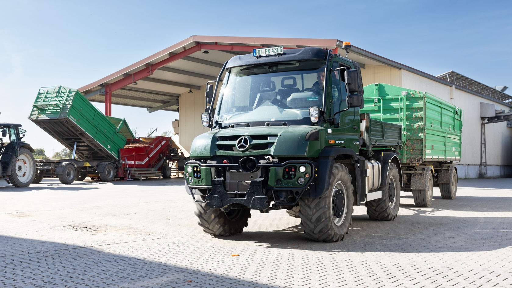 Ein grüner Mercedes-Benz Unimog steht auf einem Hof, während im Hintergrund ein Traktor mit einem Kipper Gemüse oder Erntegut entlädt.