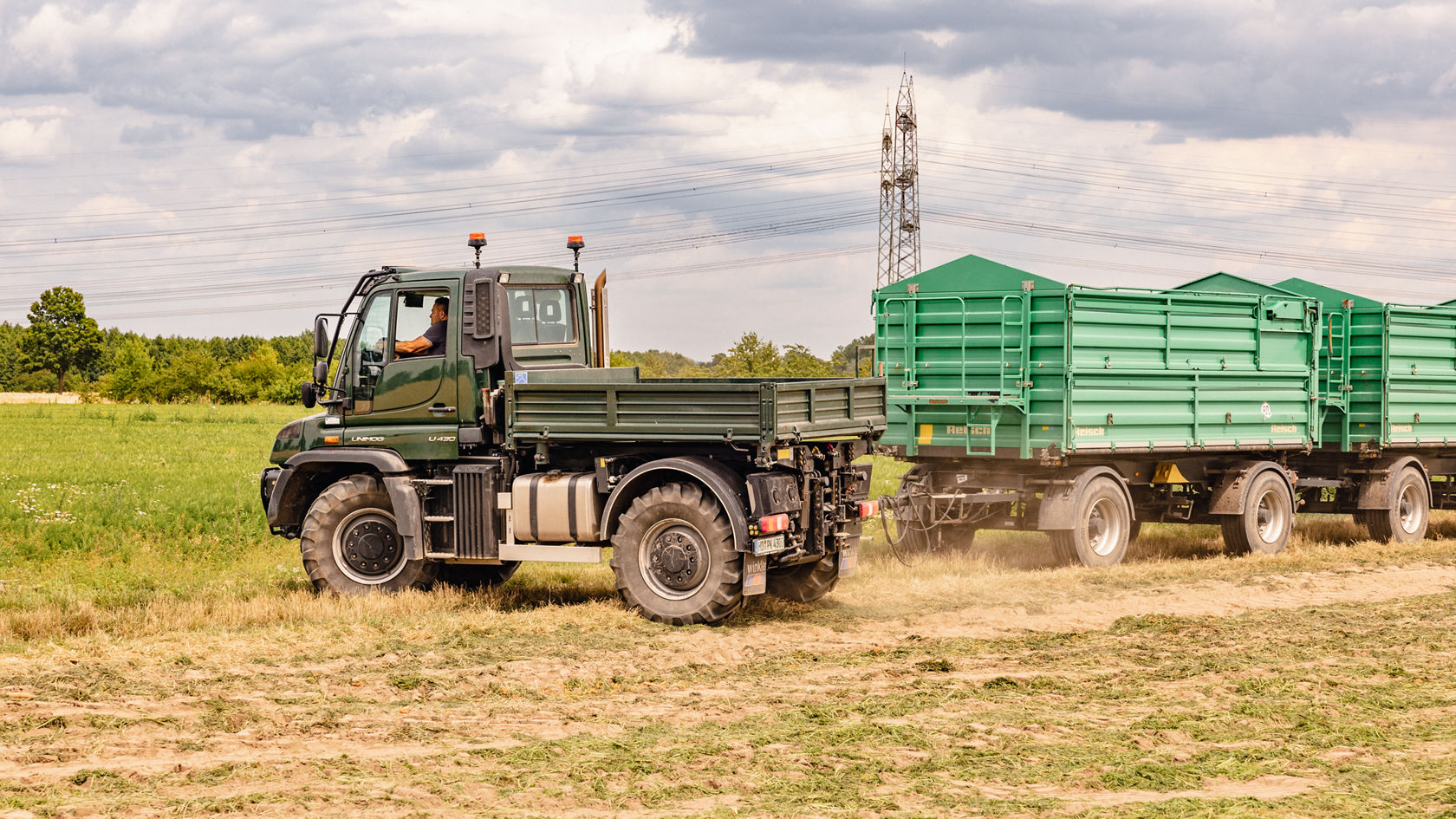 Ein Mercedes-Benz Unimog fährt über ein Feld und zieht einen Anhänger mit grünen Containern, während der Fahrer am Steuer sitzt und die Umgebung beobachtet.