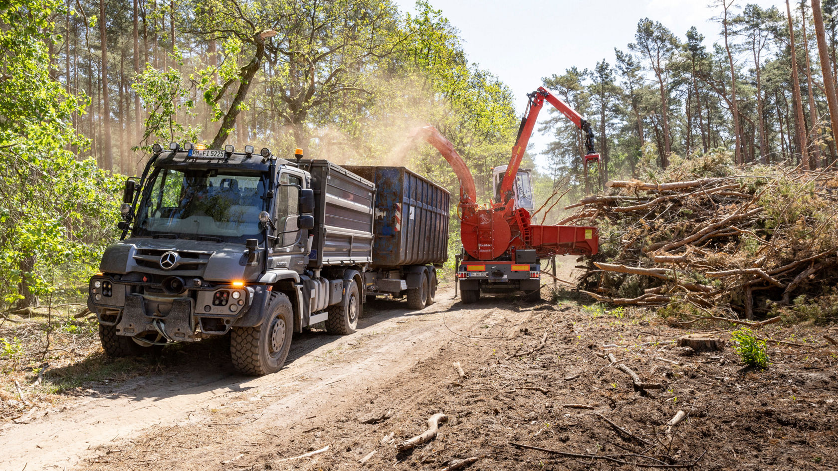 Un camion Mercedes‑Benz est stationné sur un chemin forestier, tandis qu'une déchiqueteuse de bois réduit des branches et des morceaux de bois. De la poussière s'élève dans l'air.